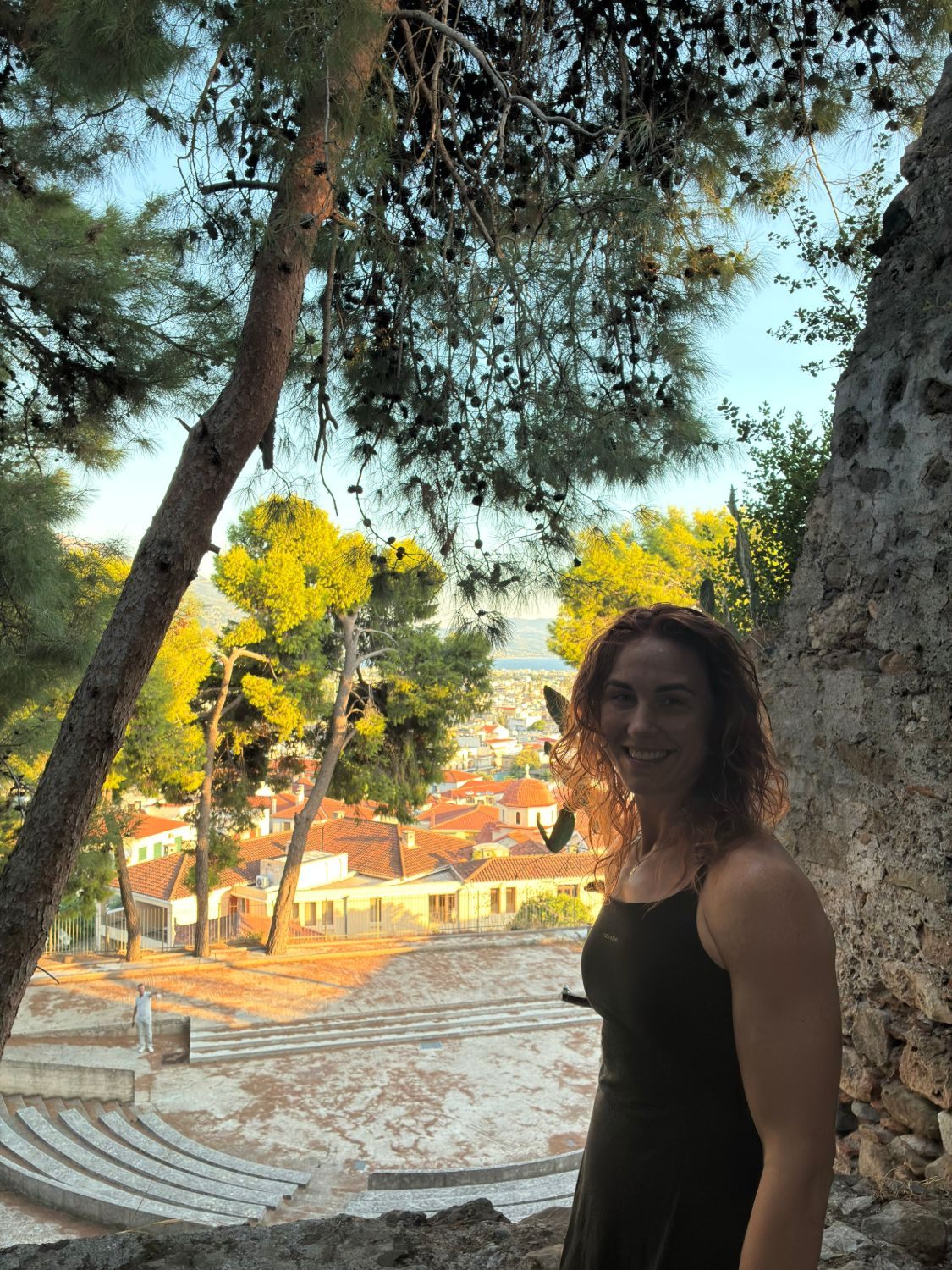 Kristine smiling under the pine trees at Kalamata Castle, with an open-air theatre and the red roofs of the city below