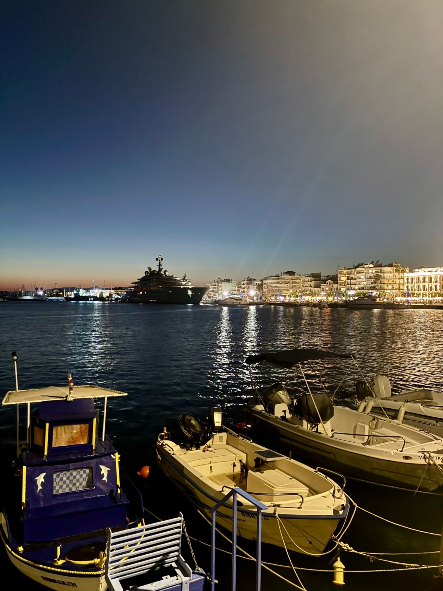 Small fishing boats in the foreground and a superyacht across the water in Kalamata harbour at dusk, with the lit-up seafront behind