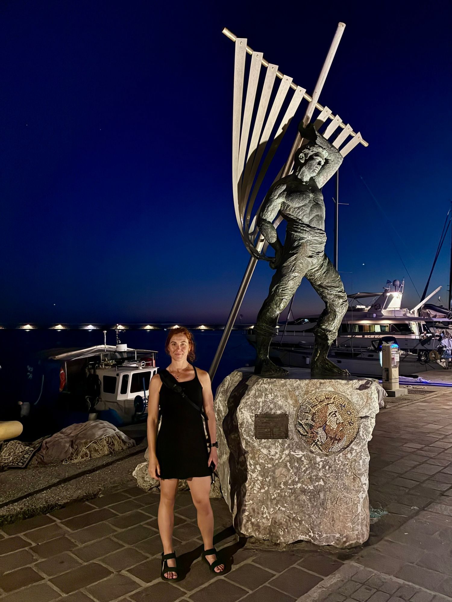 Kristine standing next to a bronze statue of a man with a sail on the harbour front in Kalamata at night