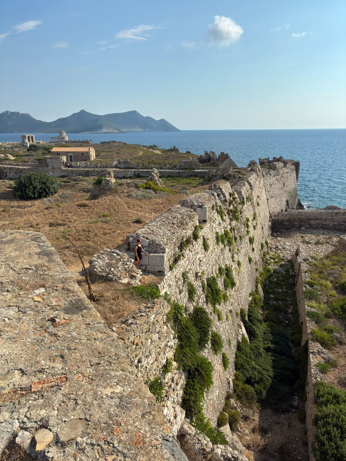 Kristine on the massive walls of Methoni Castle above a dry moat, with ruins, the Bourtzi and the sea behind
