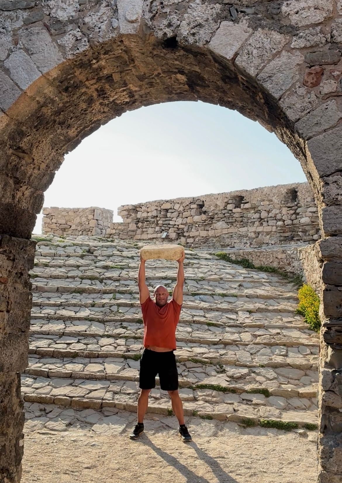 Lifting a large stone overhead framed by a stone archway inside Methoni Castle, with old stone steps behind