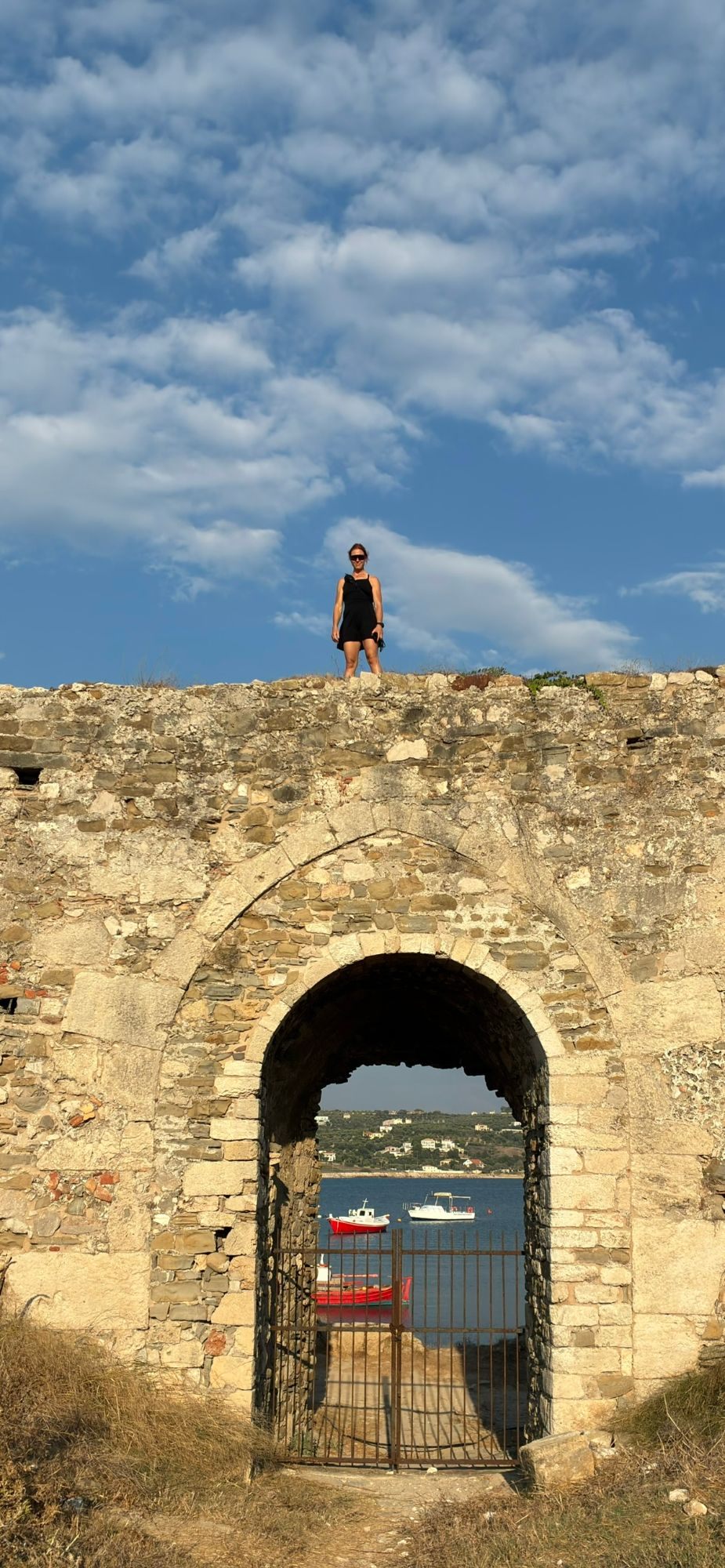 Kristine standing on top of the fortress wall at Methoni above an arched gate framing small red boats in the harbour