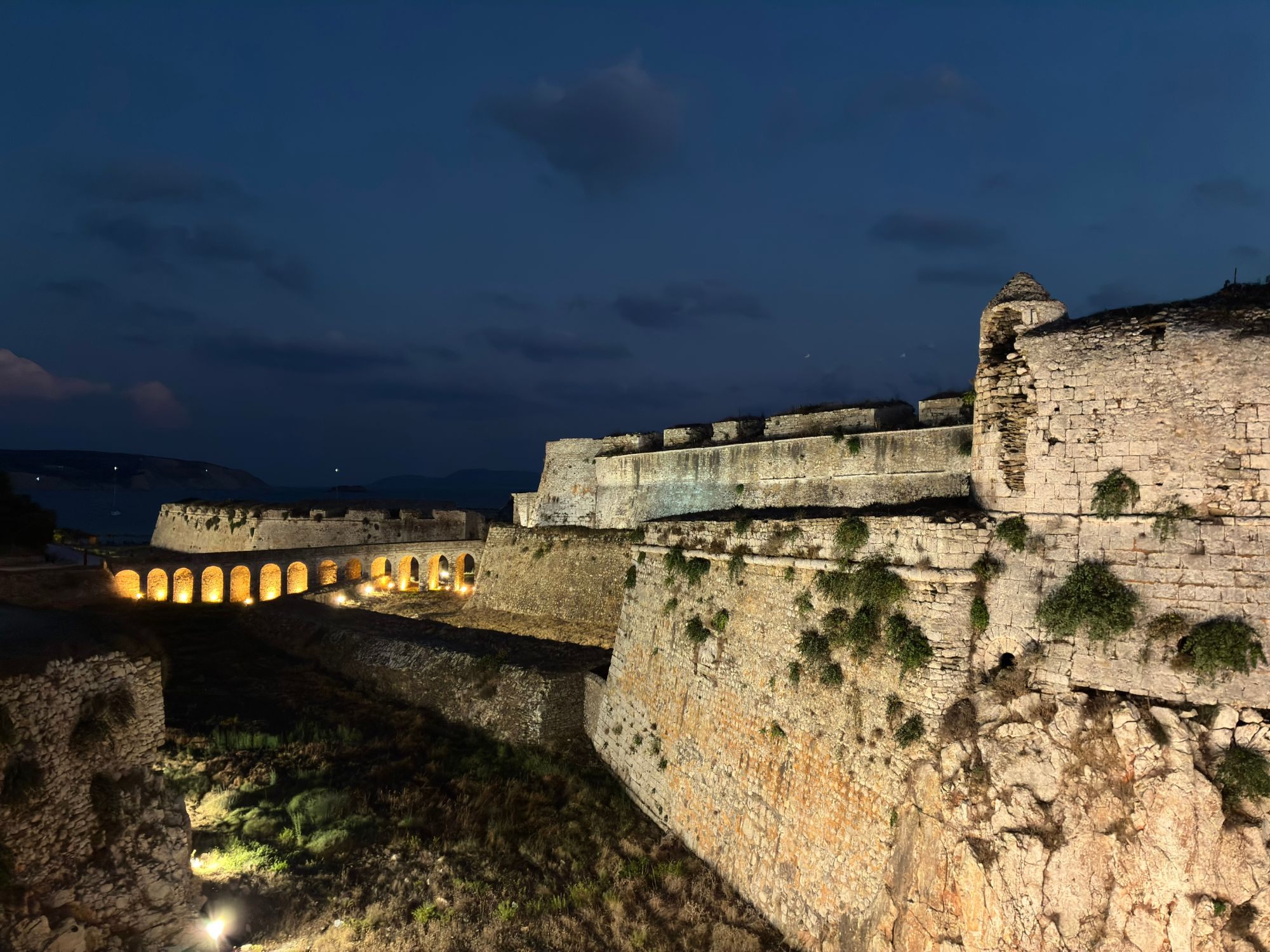 The illuminated walls and bastions of Methoni Castle at dusk, with the lit-up arches of the bridge to the entrance