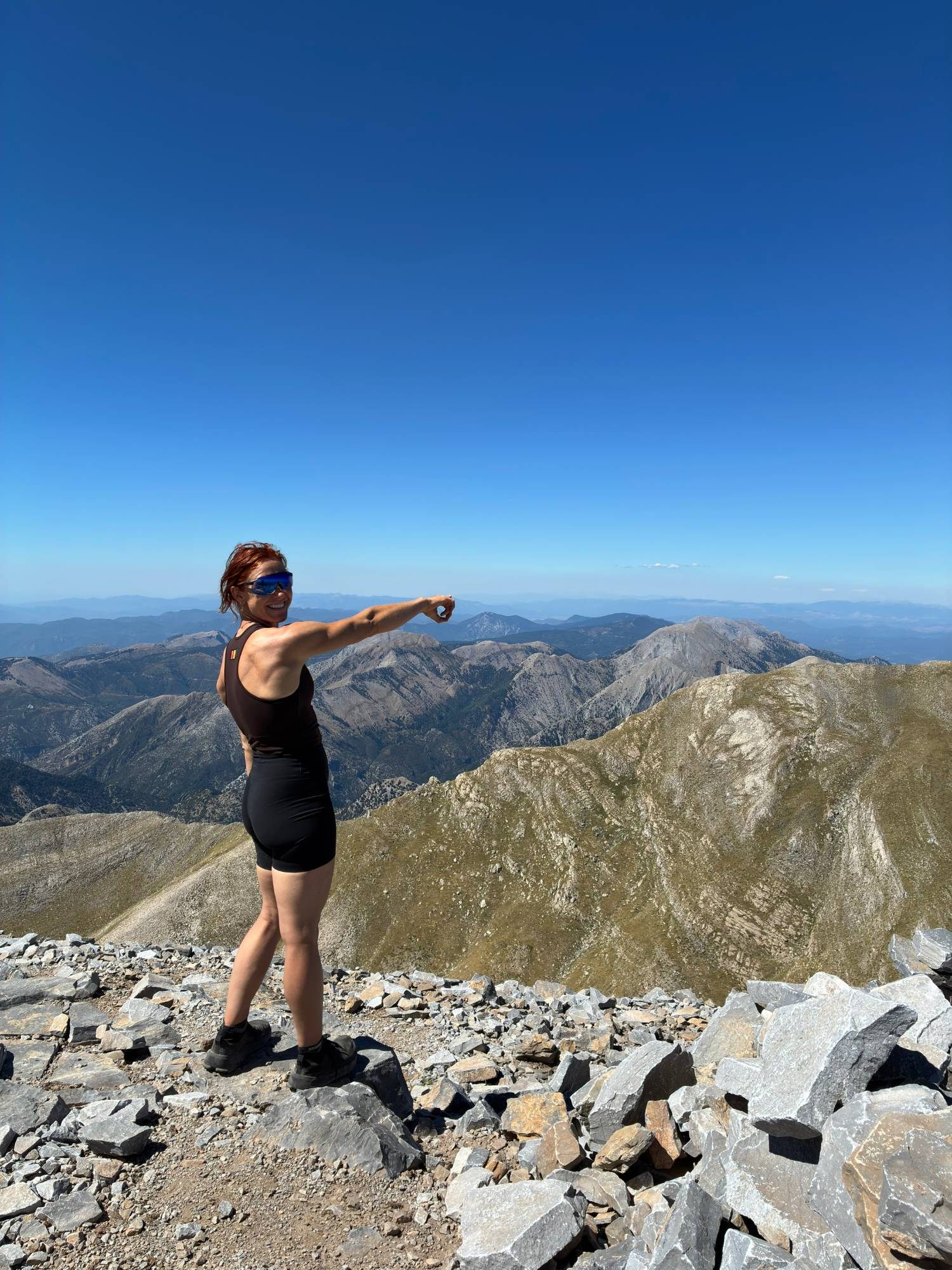 Kristine smiling and pointing out over the mountain ridges from the rocky summit of Mount Taygetos under a deep blue sky