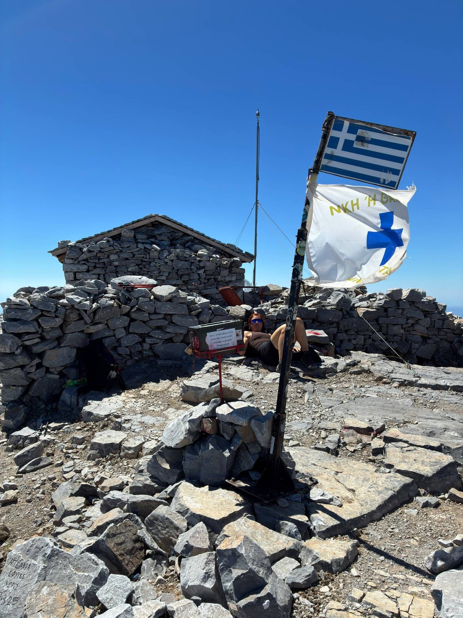 Kristine resting against the stone walls on the summit behind the Greek flag and a small message box for hikers