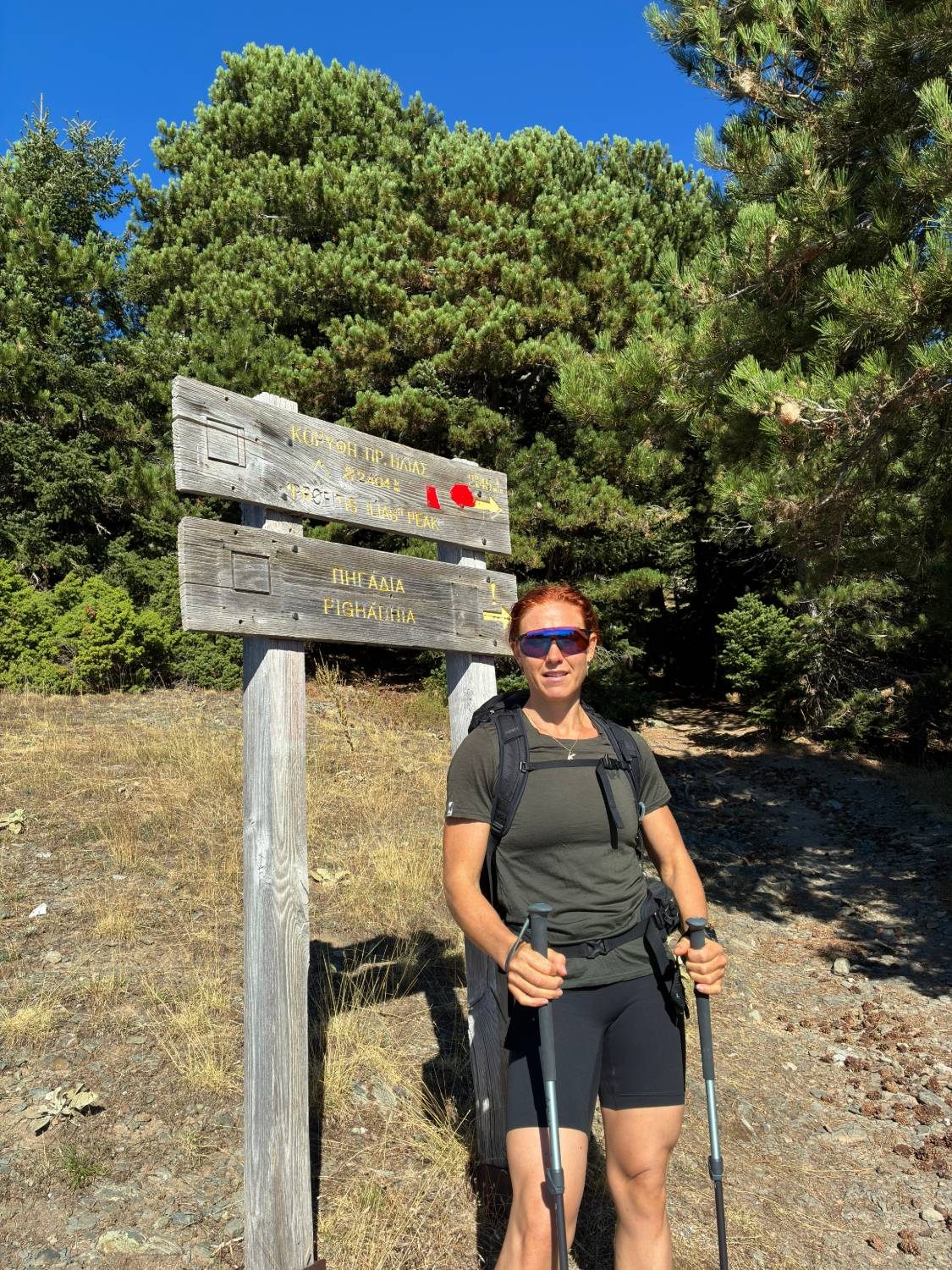Kristine with trekking poles next to the wooden signpost for Profitis Ilias Peak, 2,404 m, with red trail markings