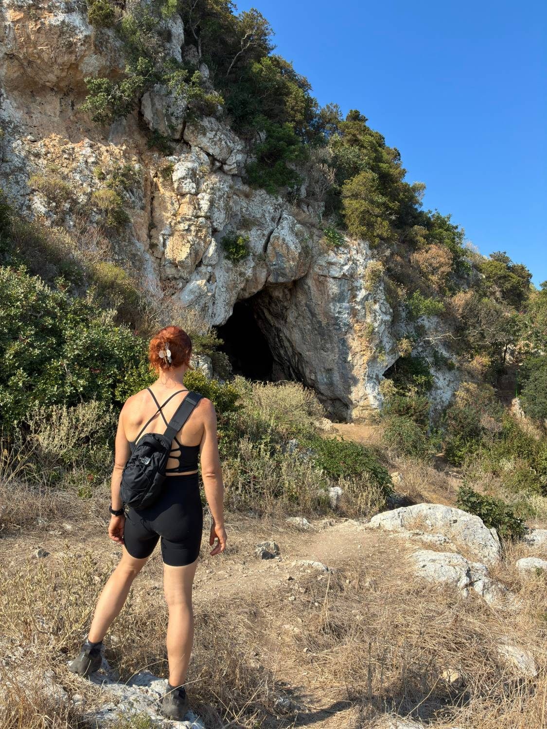 Kristine on the path below the cliff, looking up at the dark entrance of Nestor's Cave