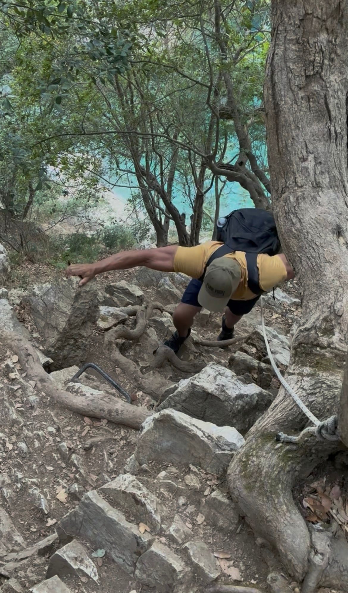 Climbing up a steep rocky section over tree roots, using a metal grip and a fixed rope, with turquoise water far below