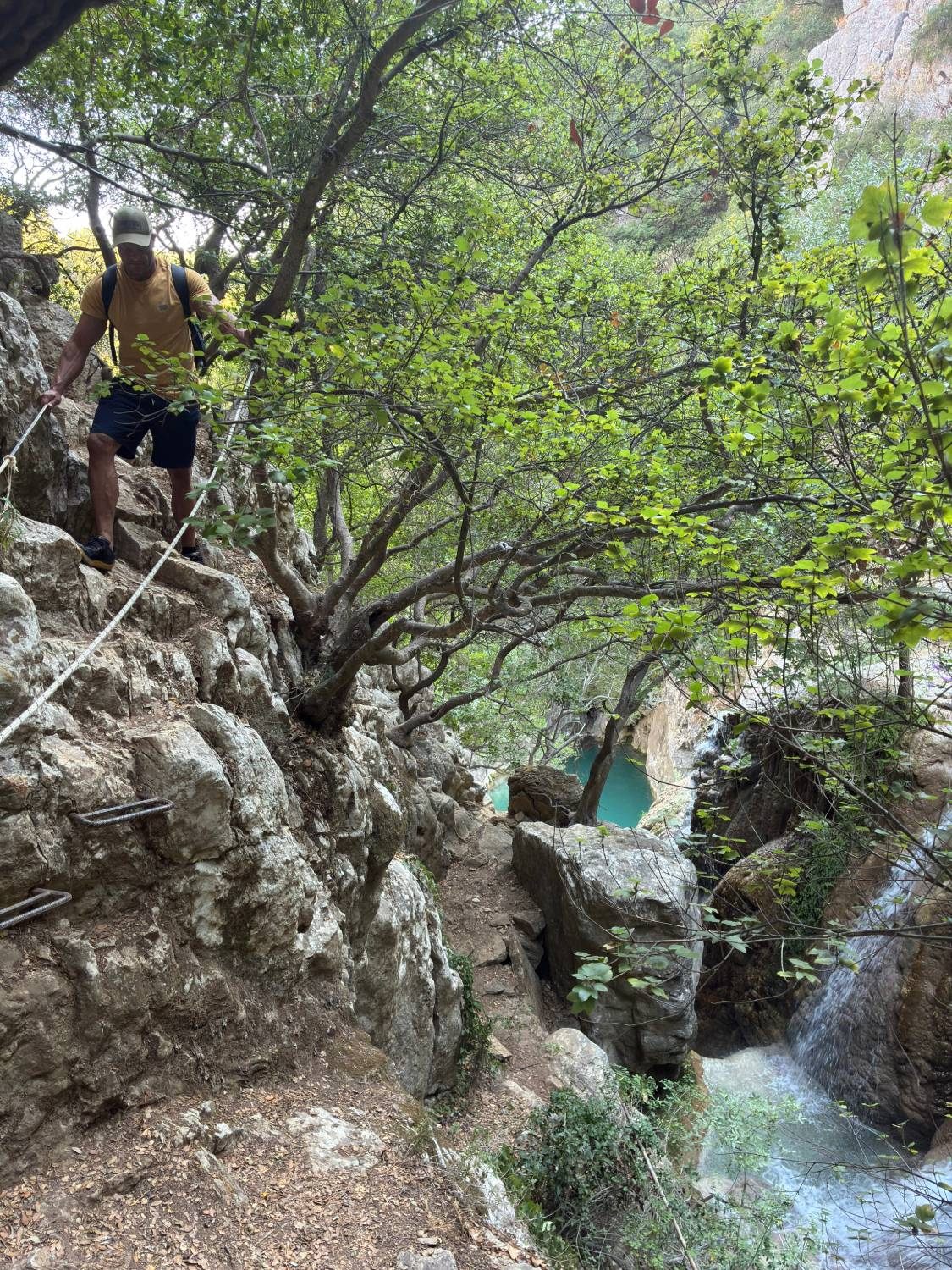 Climbing down a rocky section using a fixed rope and metal grips, with a waterfall and turquoise pool below at Polylimnio