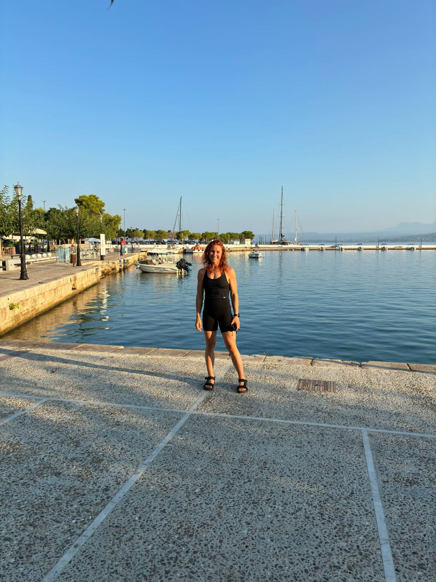 Kristine smiling on the quay in Pylos with calm blue water, small boats and sailing yachts behind